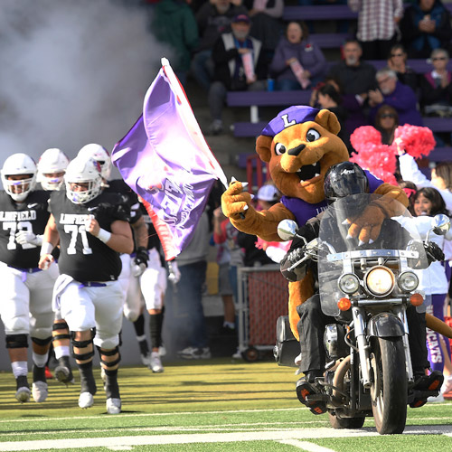 Mack riding on the back of a motorcycle as football team runs out on the field.