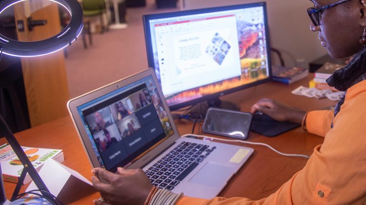 woman sits at a desk with laptop on a Zoom and second screen on a presentation